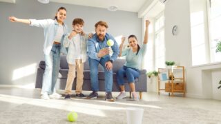 Happy family at home, mother and father with two children playing tennis ball game in living room, enjoying weekend moment, smiling parents and kids having fun together, indoor funny activity