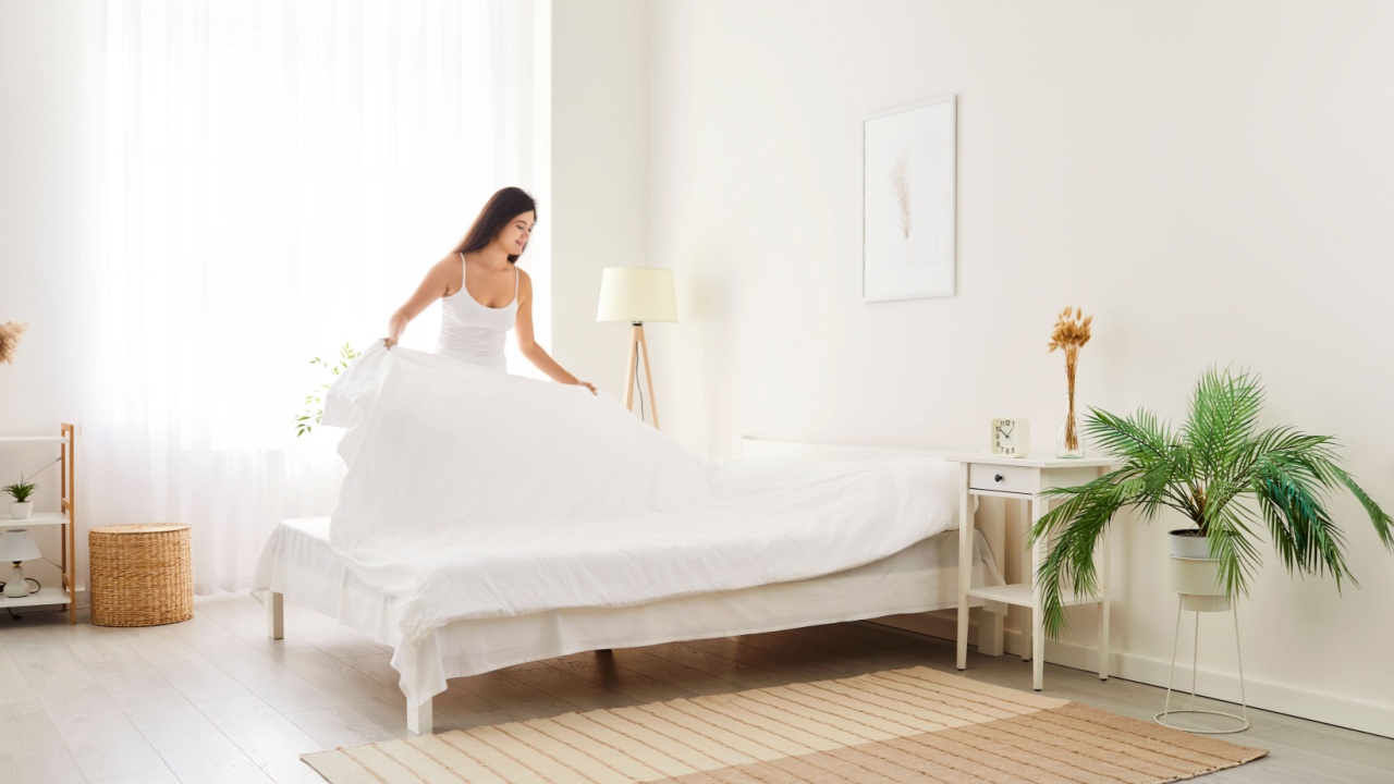 Young cheerful pretty smiling woman wearing sleepwear making her bed standing in modern cozy bedroom interior after waking up in the morning. Brunette peaceful girl making bed at home.