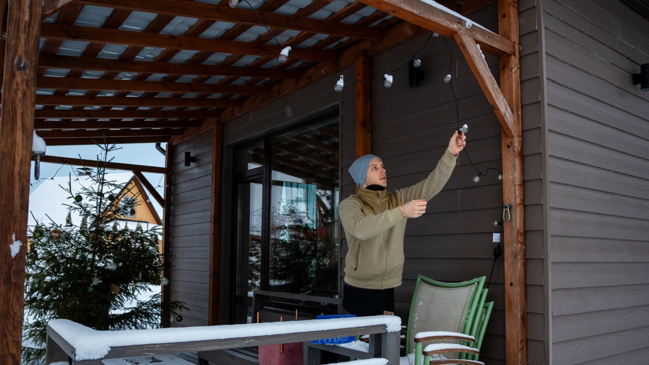 A person replaces LED string lights on a wooden terrace with a pergola roof. Snow covered trees, a house, and neighboring buildings are visible.