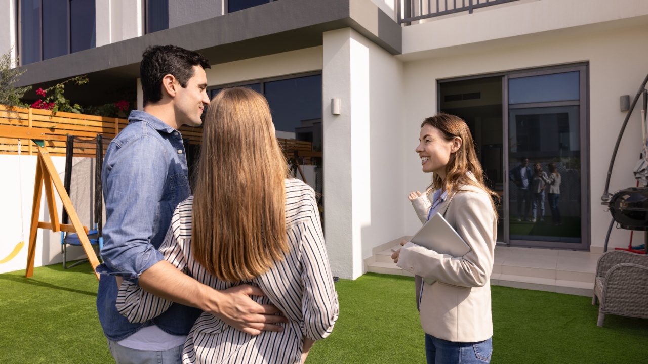 Happy real estate agent woman showing house for sale to young couple of customers, buyers, standing on yard, pointing at exterior, speaking, smiling. Renters meeting with realtor for viewing