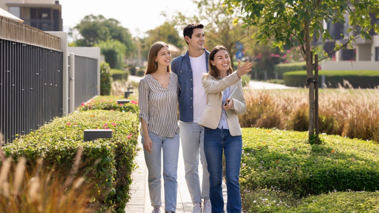 Happy young Latin couple and realtor viewing well suburban neighborhood for buying or rent house, walking along fences, greenery, pointing hand, looking away, smiling, enjoying real estate tour