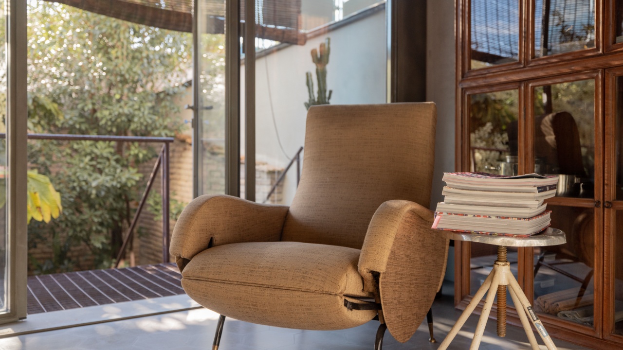 A stylish vintage armchair sits next to a small side table with stacked magazines in a bright modern interior. Sunlight streams through large glass windows, creating a warm and inviting reading nook