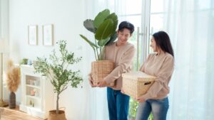 A couple joyfully holds indoor plants and wicker baskets in a bright, airy living space.