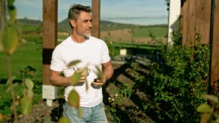 Good looking fit mid adult man standing in garden of weekend house, drinking morning coffee using smartphone. Middle aged man outdoor at home in garden, enjoying peaceful vacation and scenic view.