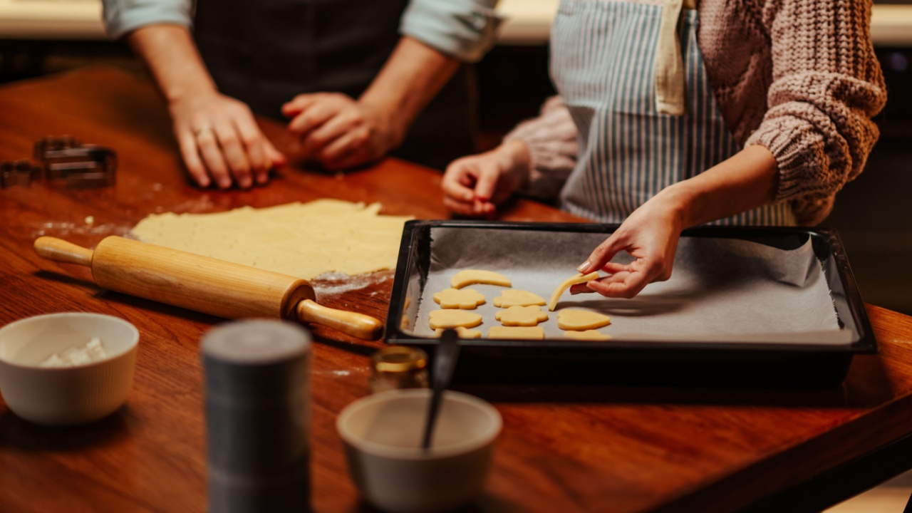 Two people prepare homemade cookies, rolling dough and placing shapes on a baking tray. The scene captures a warm, inviting kitchen atmosphere with baking tools and ingredients