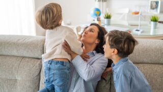 Mother is holding her youngest son, playing with him on the sofa, while her older son is sitting next to them, enjoying the family moment in their living room