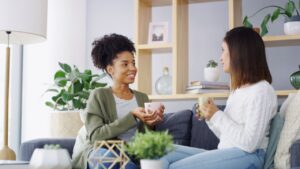 Home, coffee and women on sofa, talking and bonding together in living room. People, relax and happy friends with cappuccino, gossip and support with comfort, share secret and weekend break in lounge