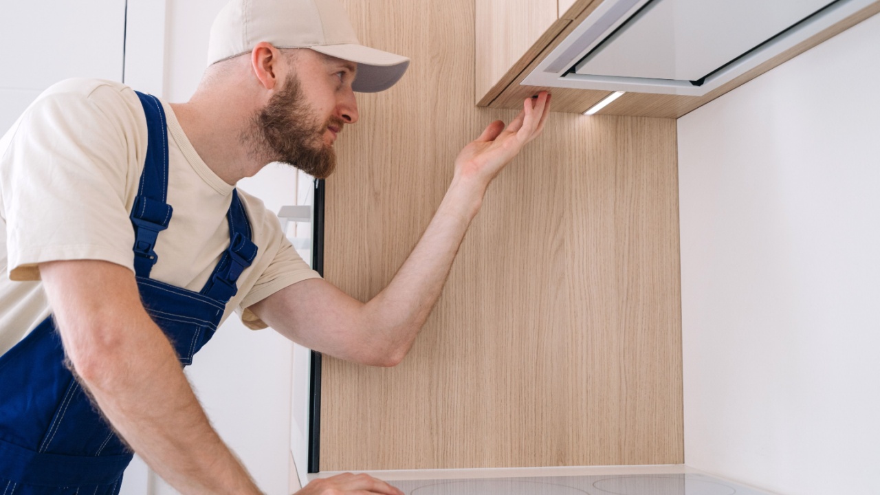 Warranty service concepts. Repairman is focused on adjusting a newly installed kitchen cabinet with led light and new cooker hood built in furniture. Residential apartment with household appliances