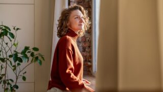 Positive thoughtful woman enjoys tranquil break at home, leaning by window feeling gentle sunlight on skin. Carefree young female with peaceful heart, joyful mind relishes beauty of day taking pause.