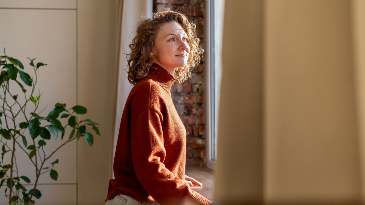 Positive thoughtful woman enjoys tranquil break at home, leaning by window feeling gentle sunlight on skin. Carefree young female with peaceful heart, joyful mind relishes beauty of day taking pause.