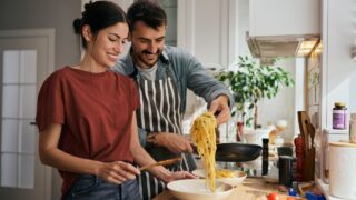 Happy man serving his girlfriend spaghetti while cooking in the kitchen.