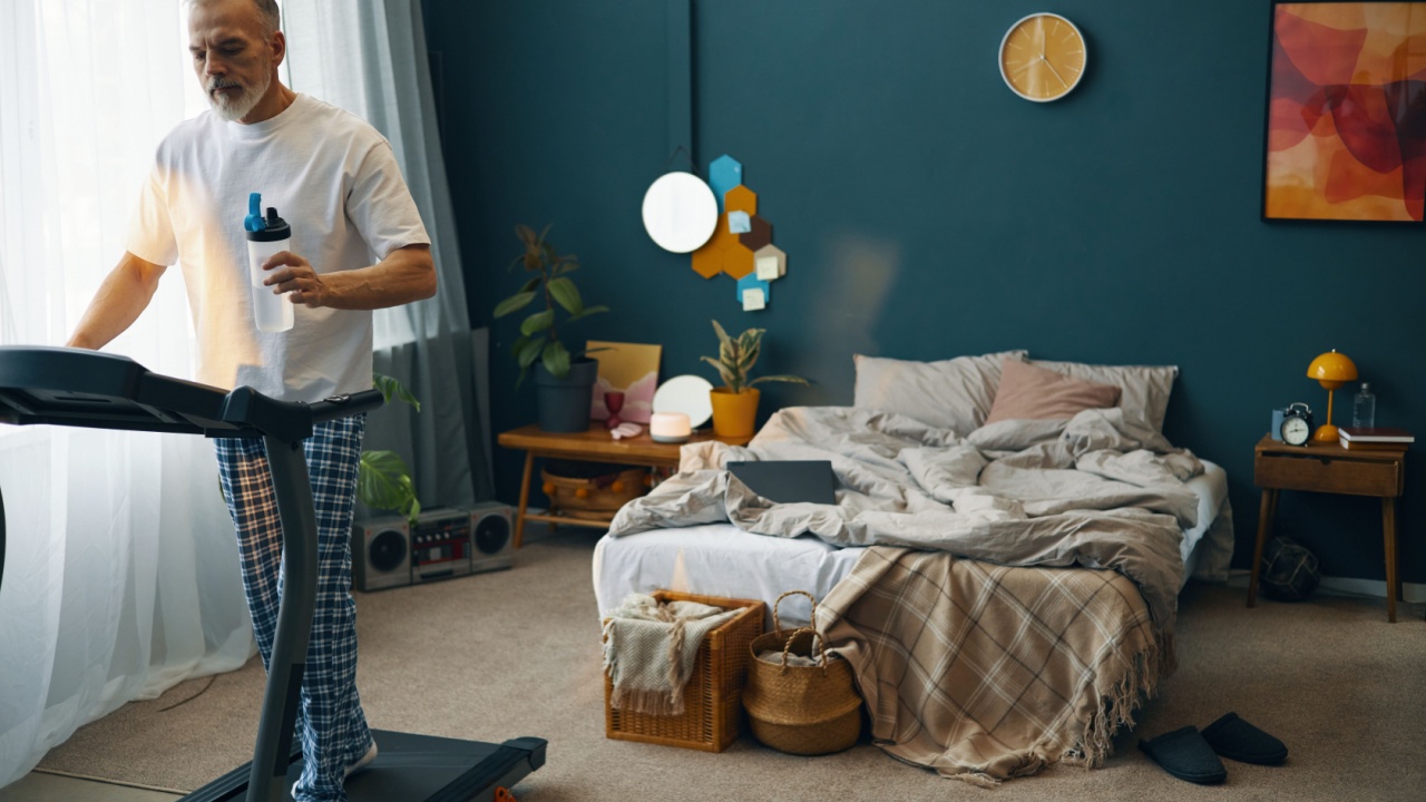 Elderly man walking on treadmill in cozy and colorful bedroom, holding a water bottle for hydration. Bedroom features tidy space with modern decor and natural light