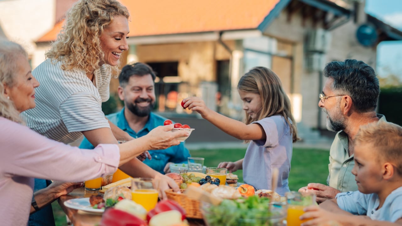 Extended family, including grandparents, parents, and children, are gathering around a table laden with fresh food, sharing a meal together in their backyard, creating a warm and joyful atmosphere
