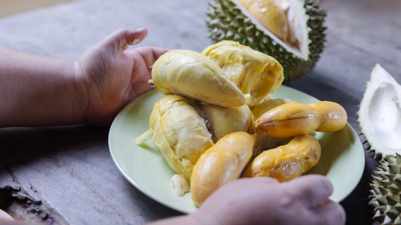 A plate full of ripe, golden-yellow durian pods is placed on a rustic wooden table, showcasing the high-sugar, creamy fruit. Hands gently hold the plate, ready to enjoy this tropical delicacy.