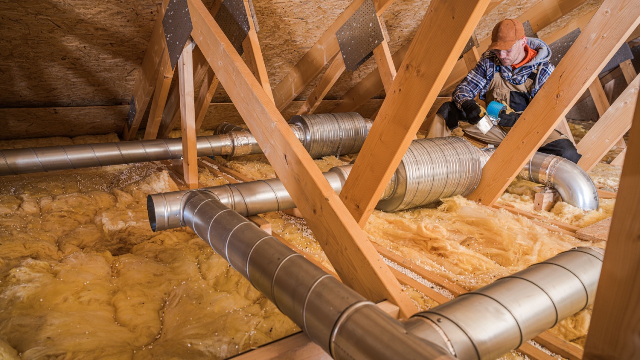A worker is fixing the ventilation ducts in an attic filled with insulation material. The setting is a residential home during the cold winter months.
