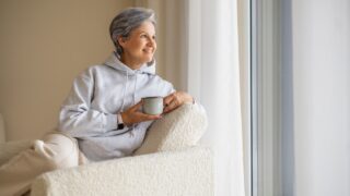Senior woman relaxing at home with cup of coffee near window in cozy hoodie and soft armchair..