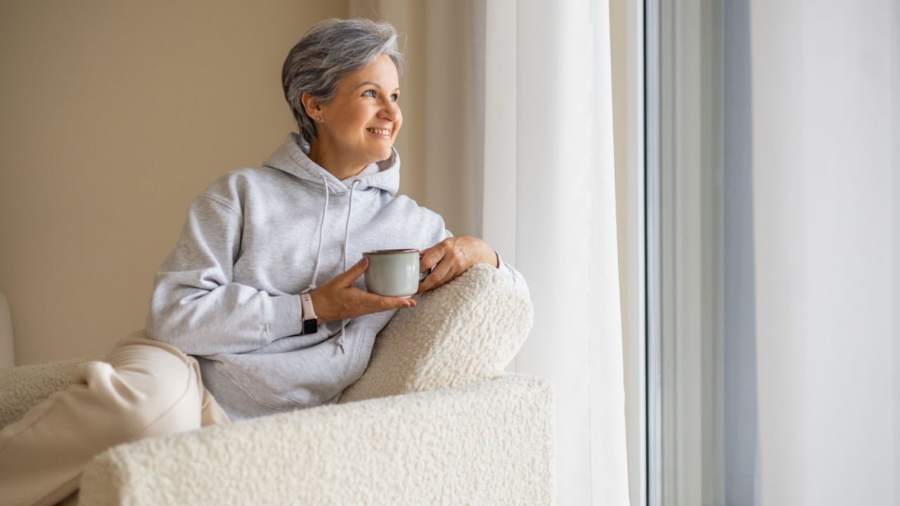 Senior woman relaxing at home with cup of coffee near window in cozy hoodie and soft armchair..