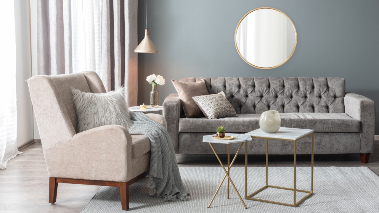 Elegant living room with tufted grey velvet sofa, beige wingback armchair, marble and gold nesting tables, round brass wall mirror, grey throw blanket, neutral rug and soft curtain lighting.