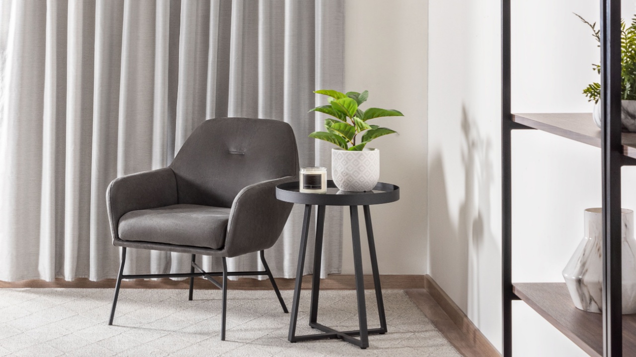 Modern Scandinavian reading corner with gray velvet armchair, round black side table, indoor plant in ceramic pot, soft textured carpet, light curtains and minimalist decorative shelving unit.