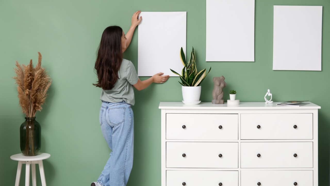 Young woman hanging empty canvas on green wall at home. Back view