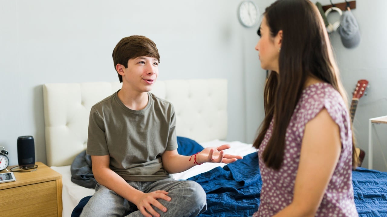 Hispanic teen boy having a conversation with his mom while sitting in his bedroom and gesturing with his hands