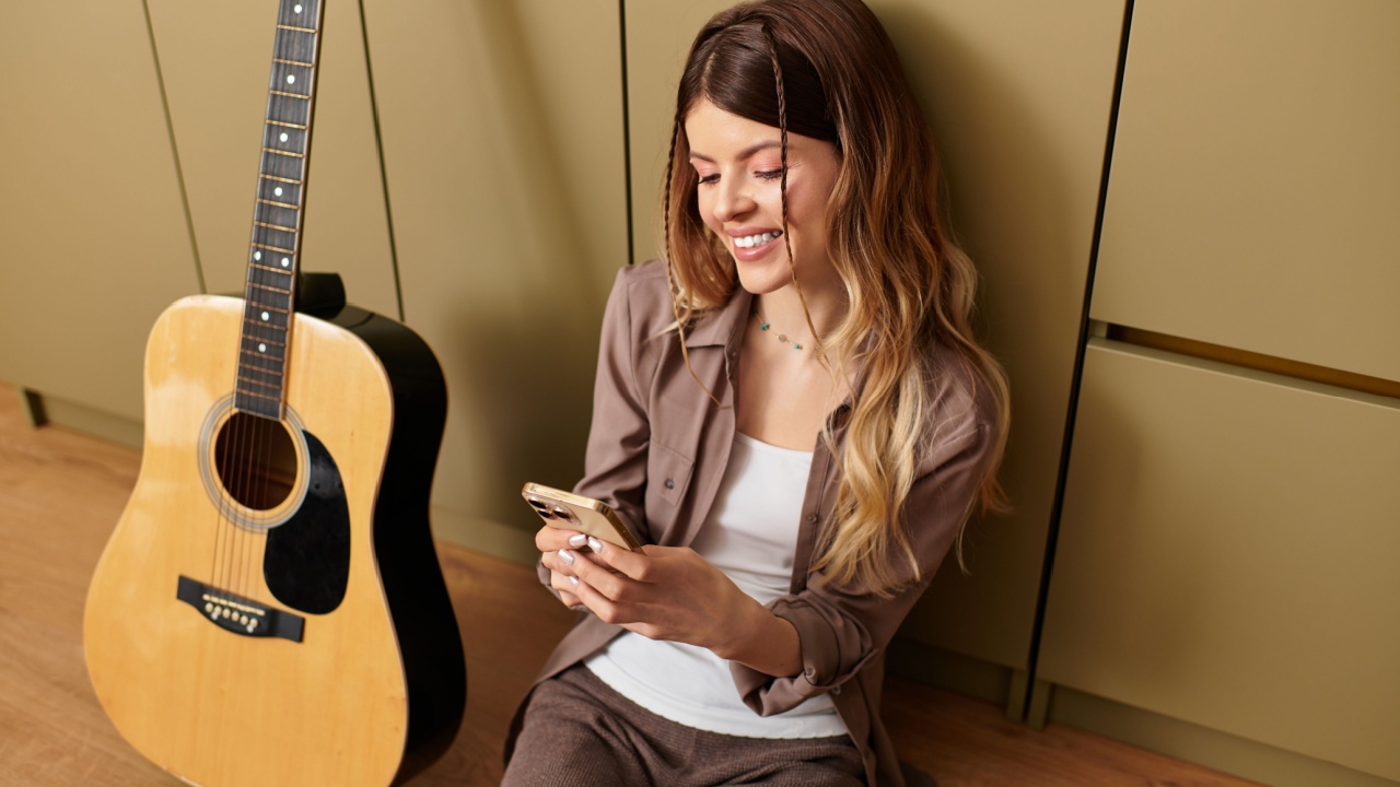 A beautiful young woman sits on the floor with a guitar, smiling at her phone in a cozy setting.