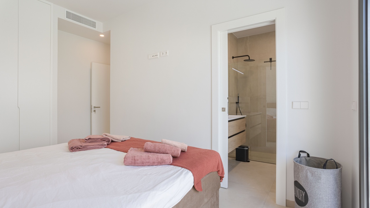 Torrevieja, Spain 15 August 2025: Bright bedroom with white walls, soft pink towels, and direct access to a modern ensuite bathroom featuring walk-in shower.