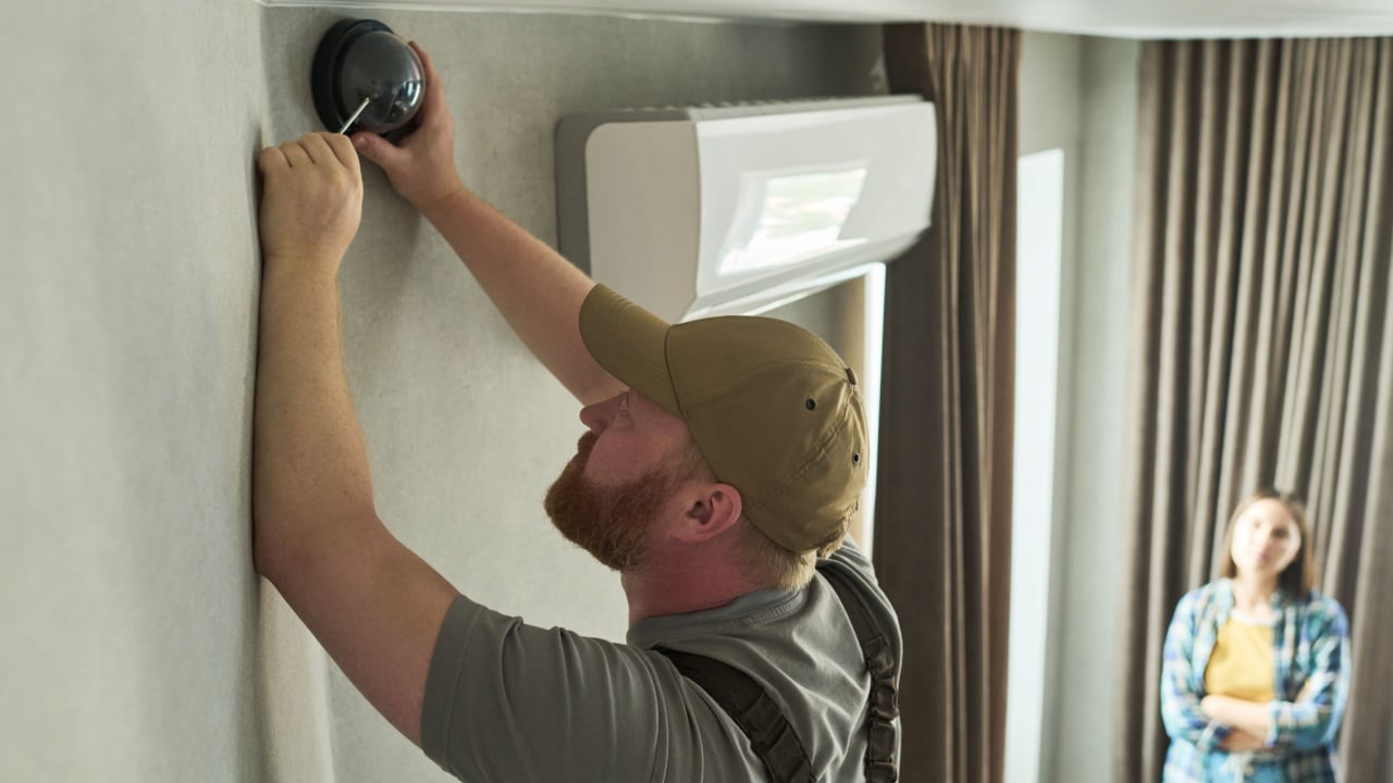 Caucasian middle aged man installing security camera on wall while Caucasian young adult woman standing in background observing process in residential interior