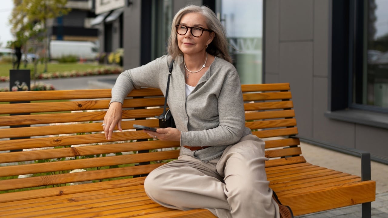 A relaxed senior woman sits on a wooden bench in an urban area, casually dressed in a sweater and slacks, focused on her smartphone while enjoying a sunny afternoon.