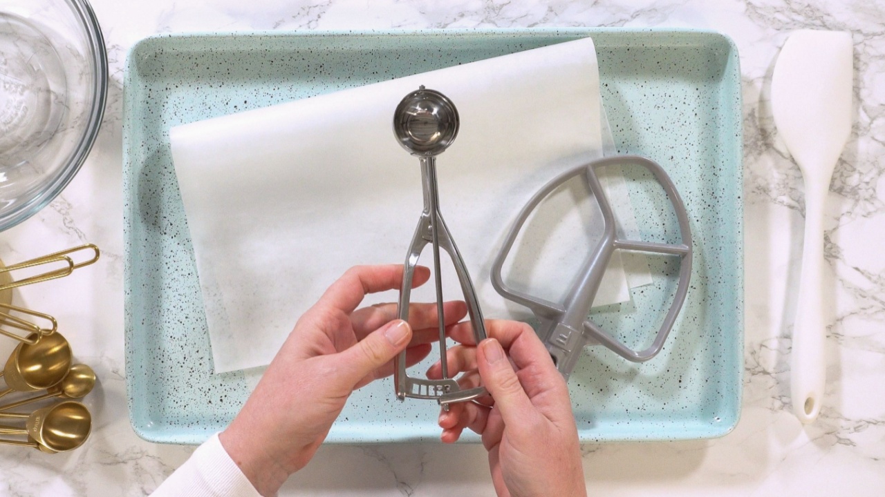Hands display a metal cookie dough scooper over a tray with parchment and baking accessories on a marble surface.