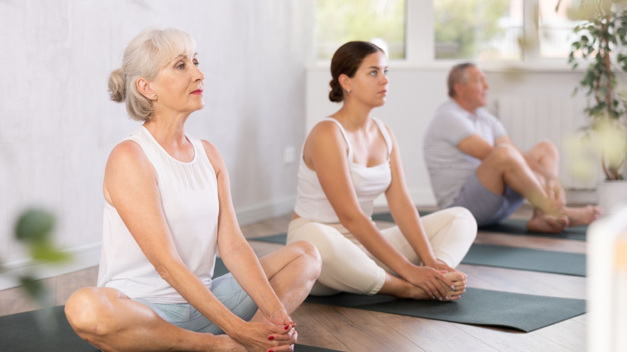 In fitness studio with small group of friends, elderly woman performs butterfly pose, Baddha Konasana. Elderly woman participates in yoga practice.Concept of active lifestyle of older generation