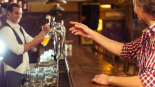Side view of man in casual clothes ordering beer while sitting at bar counter in pub, a bartender in the background