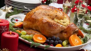 Roasted herb rubbed turkey garnished with fresh grapes, oranges, and cranberry is ready for Christmas dinner. Ornaments, Champagne, candles, and other Christmas decorations on feast table.