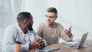 Smiling multicultural colleagues sitting at table and talking in creative agency