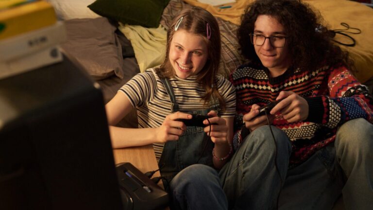 Teenage girl and boy smiling while playing video games together on bed in cozy room Portrait of young people having fun. Vintage video game