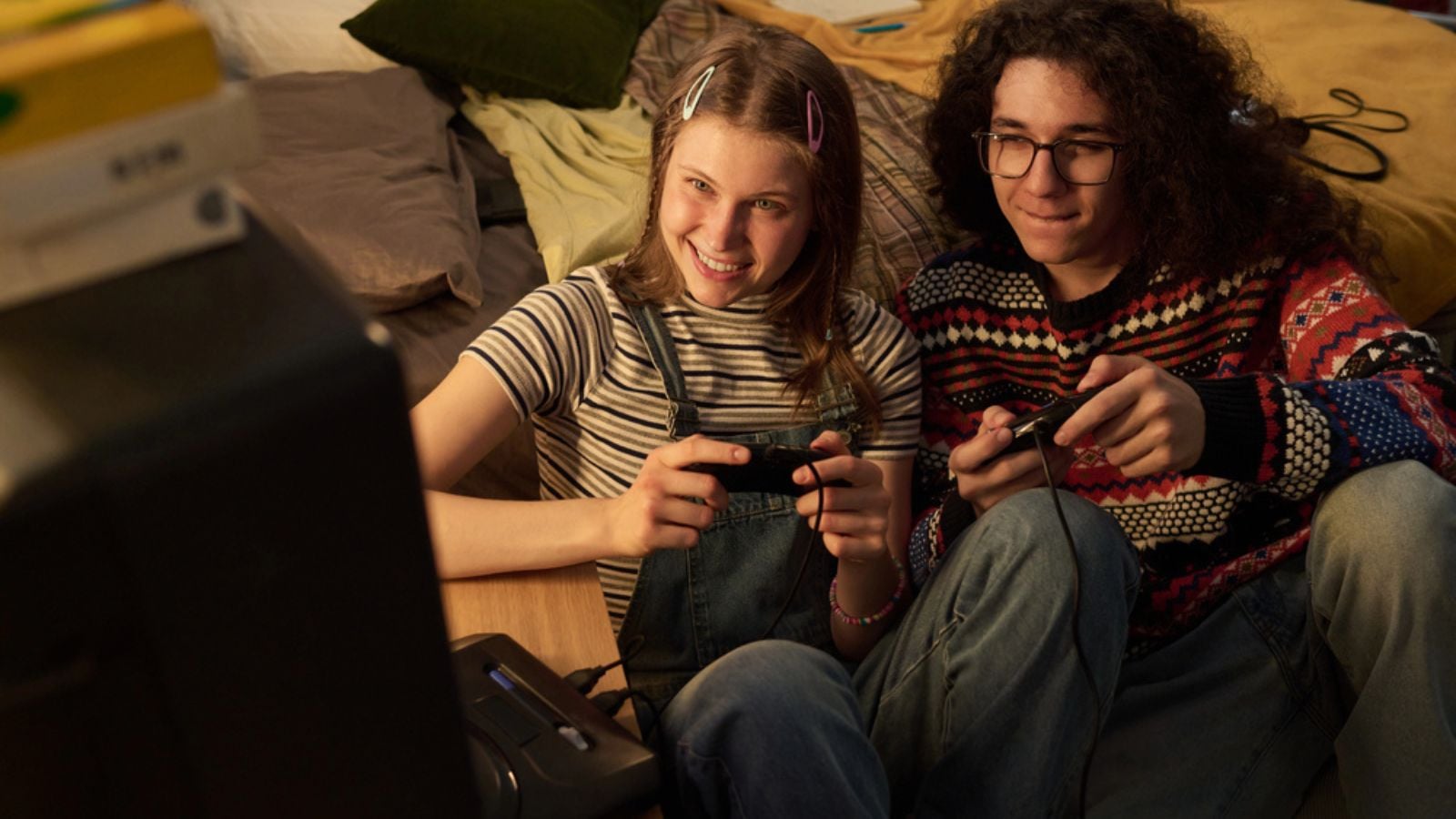 Teenage girl and boy smiling while playing video games together on bed in cozy room Portrait of young people having fun. Vintage video game