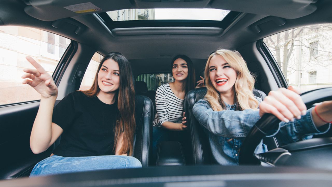 Three beautiful young cheerful women looking at pointed hand direction with smile while sitting in car
