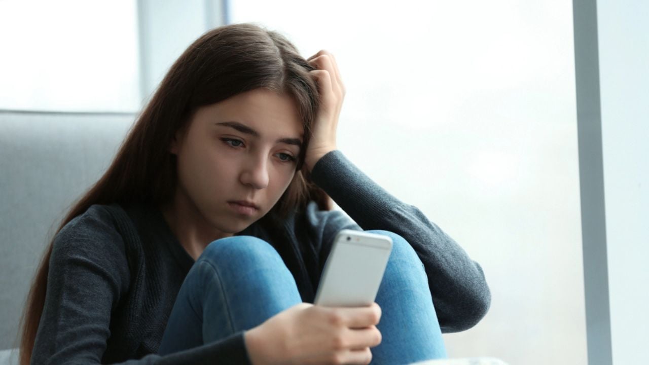 Upset teenage girl with smartphone sitting at window indoors.