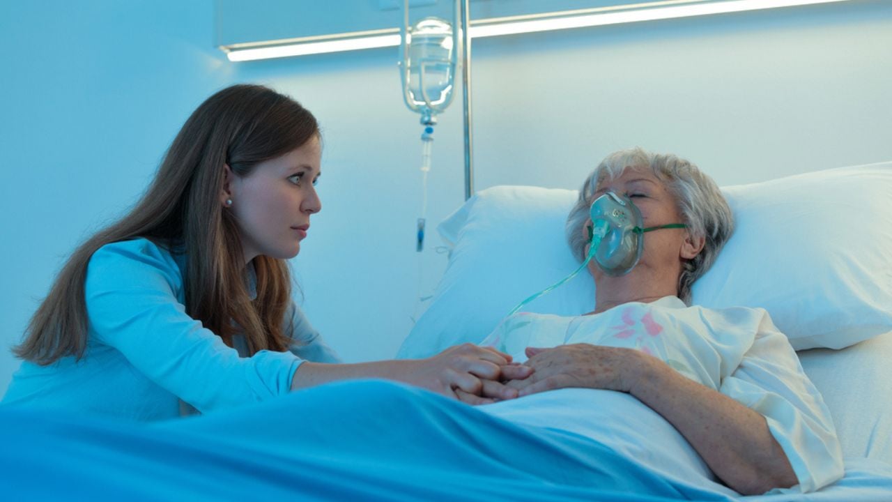 Worried daughter holding the hand of her mother in hospital as the elderly woman lies asleep or in a coma on a bed wearing a positive oxygen mask to assist breathing
