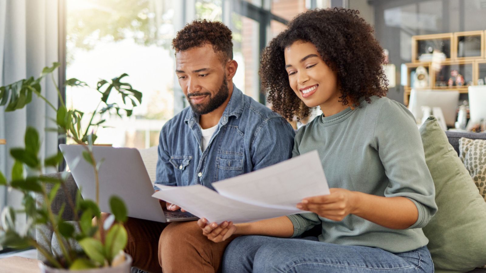 Young happy mixed race couple going through documents and using a laptop at a table together at home. Hispanic husband and wife planning and paying bills