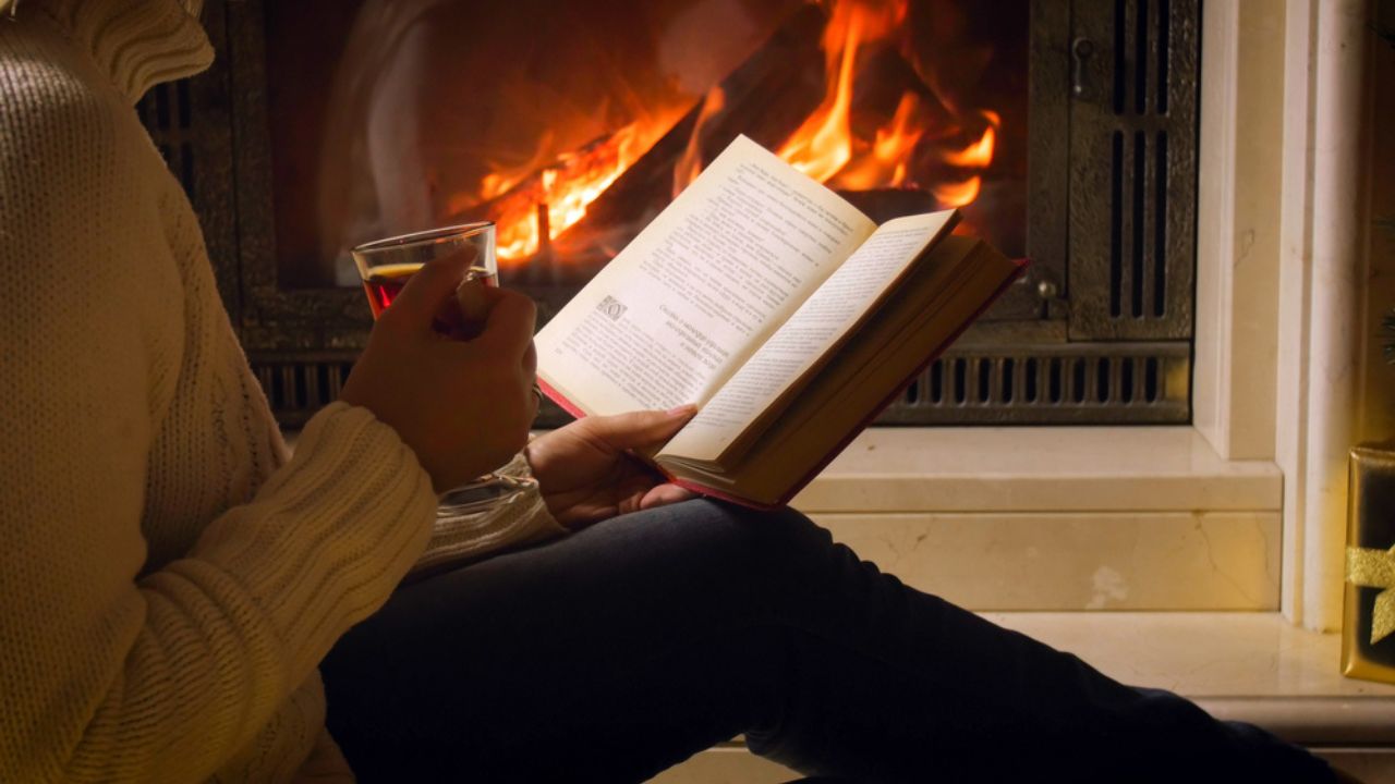 Young woman drinking tea and reading book by the fireplace at night