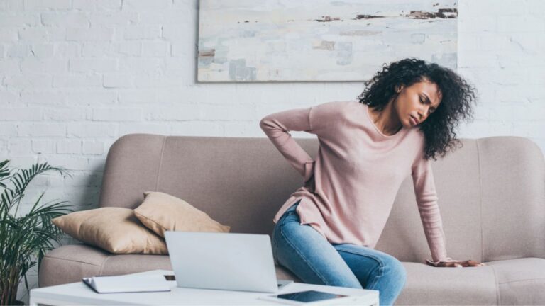 nhappy african american woman suffering from back pain while sitting on sofa near table with digital devices