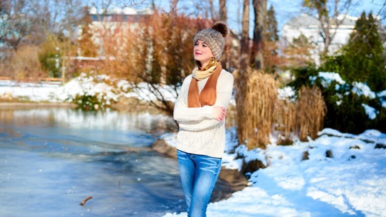 a young woman in a hat and scarf stands by a snowy pond