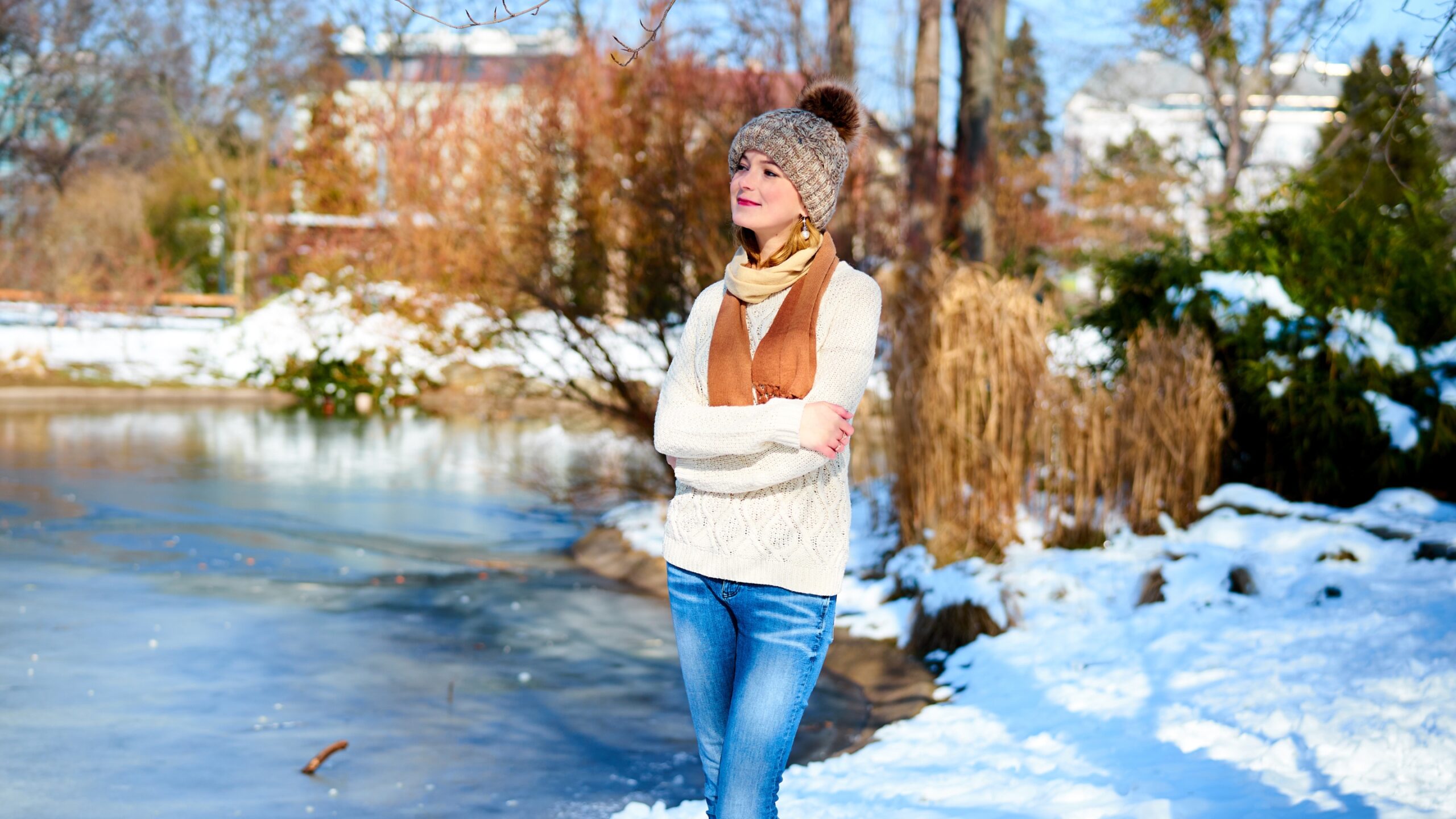a young woman in a hat and scarf stands by a snowy pond