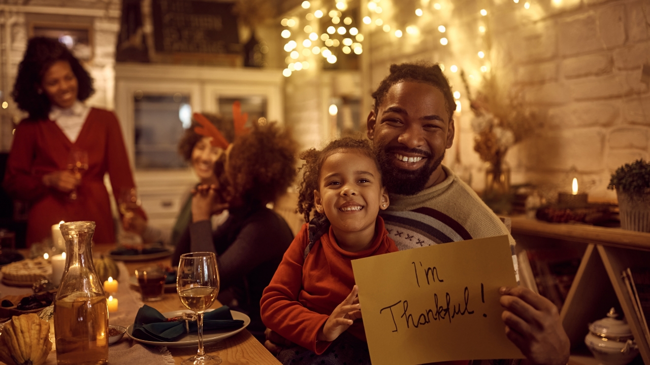 a father and little girl hold a sign that reads "I'm thankful" while sitting at dinner with family