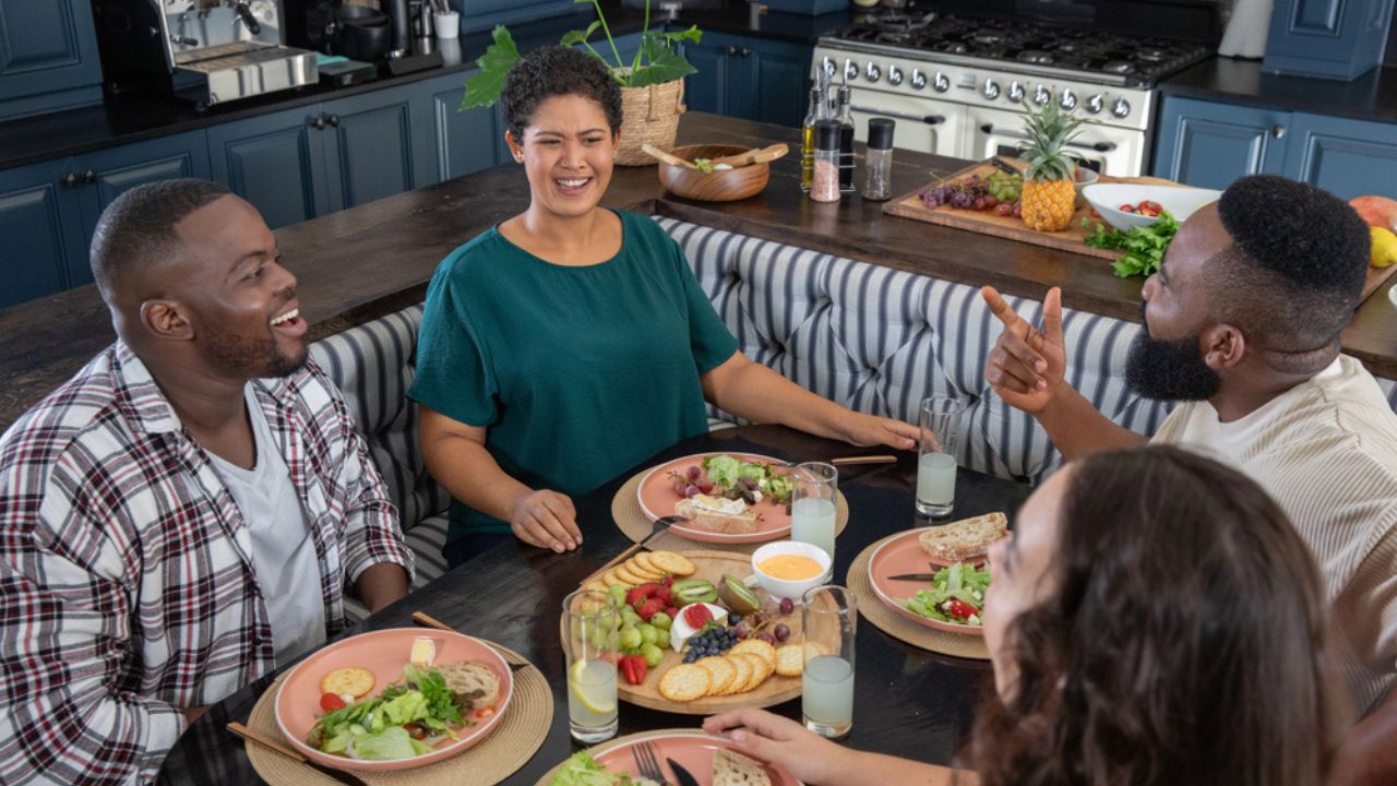 Diverse friends chatting and laughing at dining table in home kitchen with cheese platter.