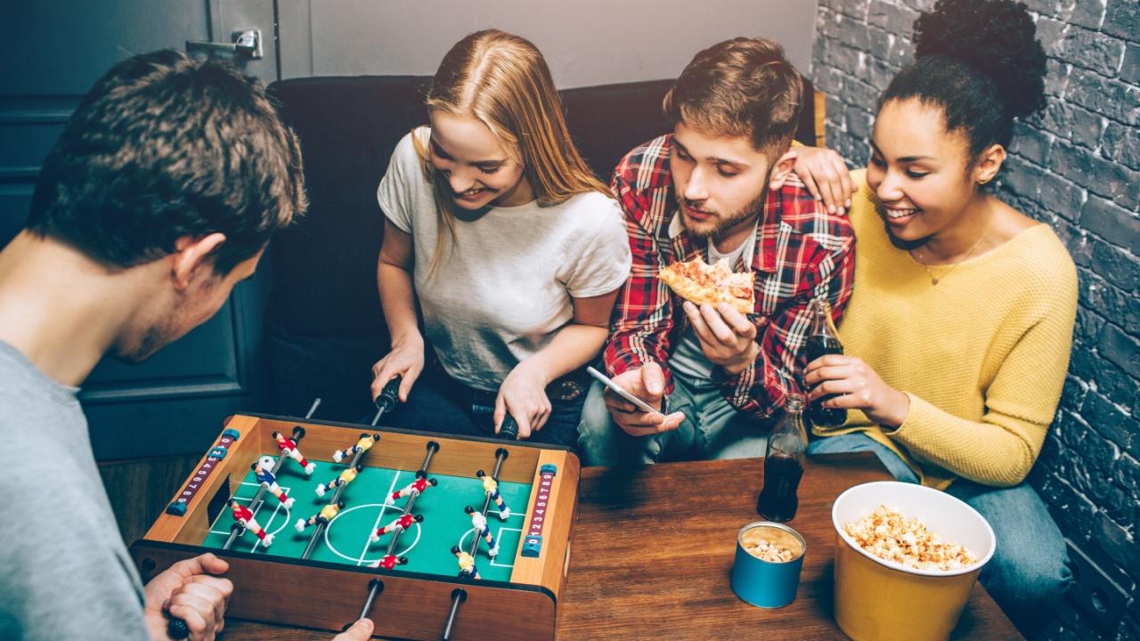 Two people are playing football board game on the table while another two are watching on it and trying to support players as best as they can. Nobody knows who will win