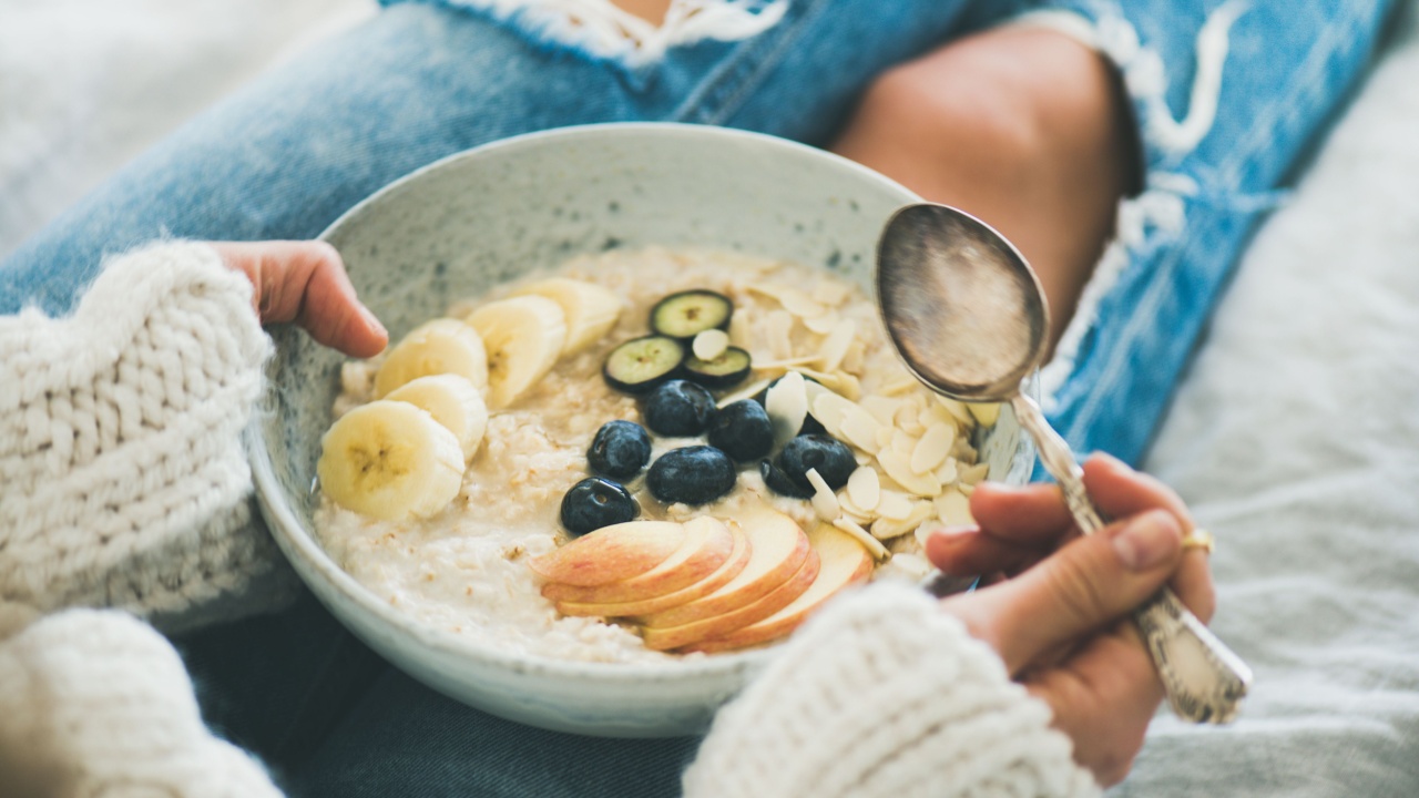 Healthy winter breakfast in bed. Woman in woolen sweater and shabby jeans eating vegan almond milk oatmeal porridge in bowl with berries, fruit and almonds. Clean eating, vegetarian food concept