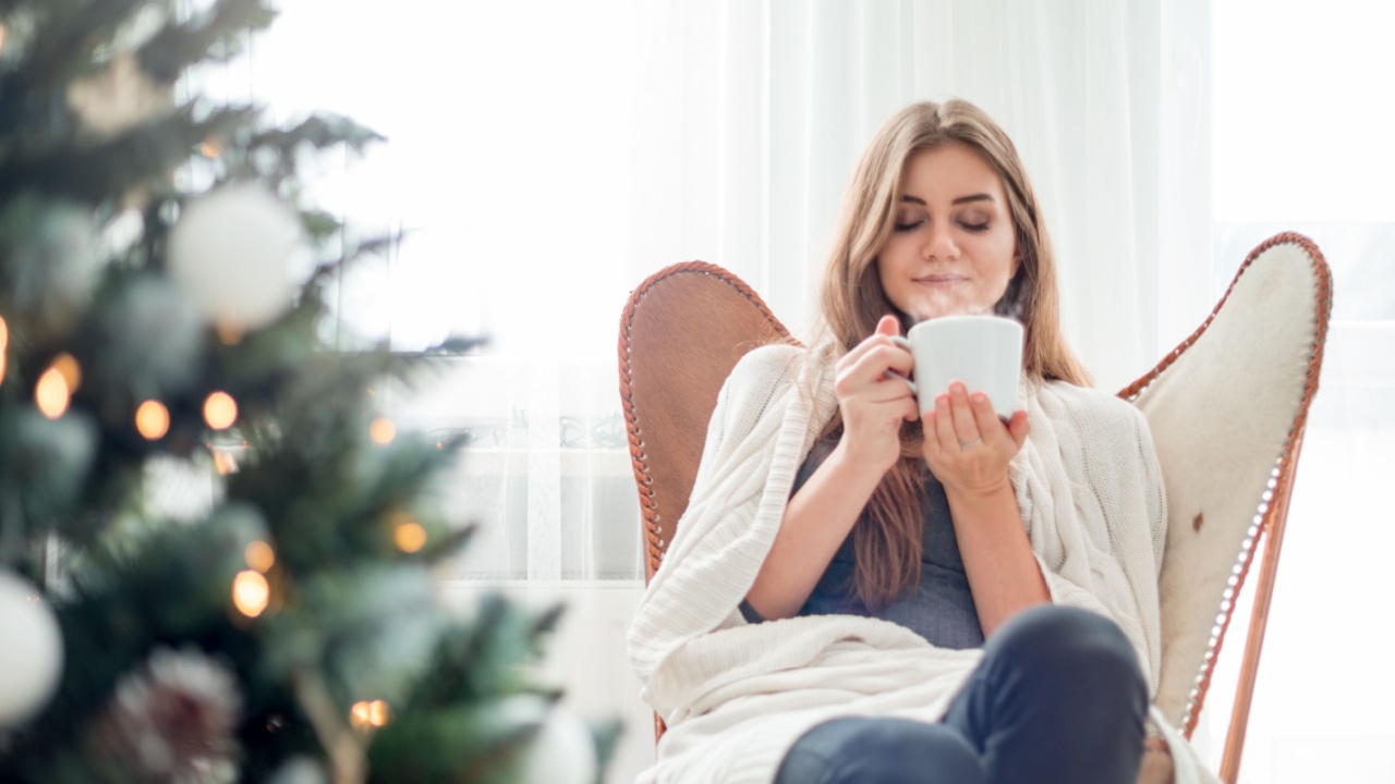 Young relaxed woman resting near Christmas tree and drinking cup of hot tea or coffee