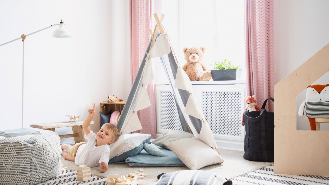 Smiling kid on the floor and plush toy in bedroom interior with pouf and tent next to lamp